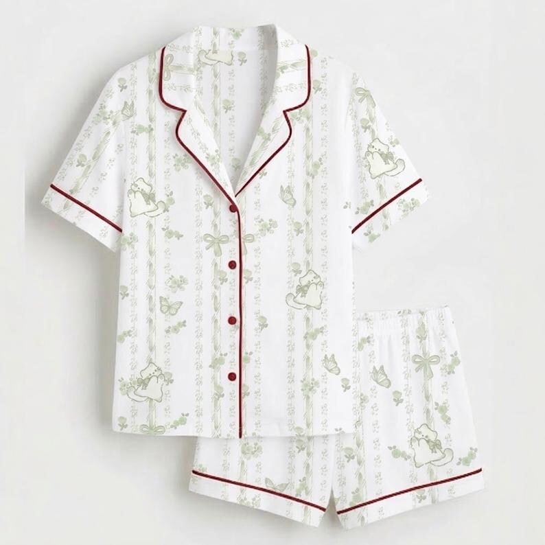 May include: White pyjama set featuring a pattern of green cats, bows, and floral designs. The short-sleeved top and shorts have burgundy trim and buttons. A comfortable sleepwear set.