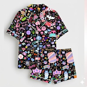 May include: Black pajama set with a vibrant print of colorful icons, text, and illustrations. The set includes a short-sleeved button-up shirt and matching shorts, both trimmed with white piping. The design features words like "Cute", "Happy", and "Beautiful".