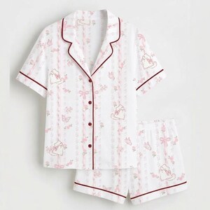 May include: White pyjama set featuring a pattern of pink bows, flowers, and cartoon cats. The short-sleeved top and shorts have burgundy trim. The top has a button-down front.