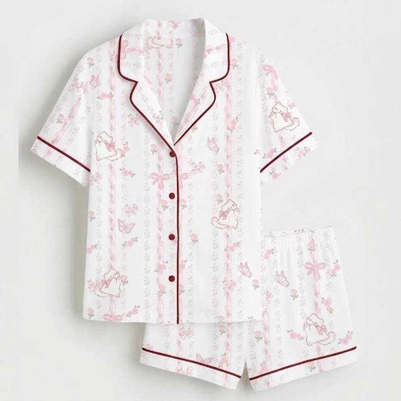 May include: White pyjama set featuring a pattern of pink bows, flowers, and cartoon cats. The short-sleeved top and shorts have burgundy trim. The top has a button-down front.