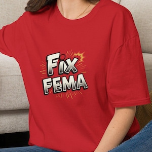 Könnte beinhalten: Ein rotes kurzärmeliges T-Shirt mit einem Grafikdesign auf der Brust, das den Text "FIX FEMA" in einer fetten Schrift im Comic-Stil zeigt, akzentuiert durch ein gelb-orangefarbenes Sternen-Hintergrundelement, kombiniert mit einer blauen Jeans.