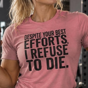 May include: A dusty rose short-sleeved t-shirt featuring the black printed text "DESPITE YOUR BEST EFFORTS, I REFUSE TO DIE." The shirt has a crew neckline and is paired with black athletic shorts that feature a small white logo on the left leg.