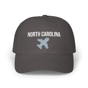 Embroidered dad cap with the words “NORTH CAROLINA” above a small light blue airplane graphic centered on the front; one-size-fits-all cotton twill hat with a curved visor and adjustable strap, available in the same color chart options as the other state caps (pink, white, black, light blue, khaki, stone, green camo, dark grey, spruce, and navy).
