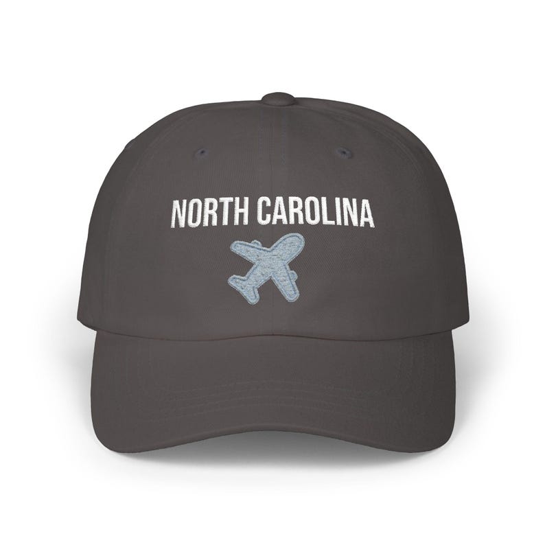 Embroidered dad cap with the words “NORTH CAROLINA” above a small light blue airplane graphic centered on the front; one-size-fits-all cotton twill hat with a curved visor and adjustable strap, available in the same color chart options as the other state caps (pink, white, black, light blue, khaki, stone, green camo, dark grey, spruce, and navy).