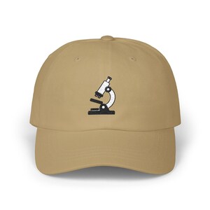May include: A tan baseball cap with a black and white embroidered microscope design on the front. The cap has a curved brim and a classic six-panel construction, suitable for casual wear.