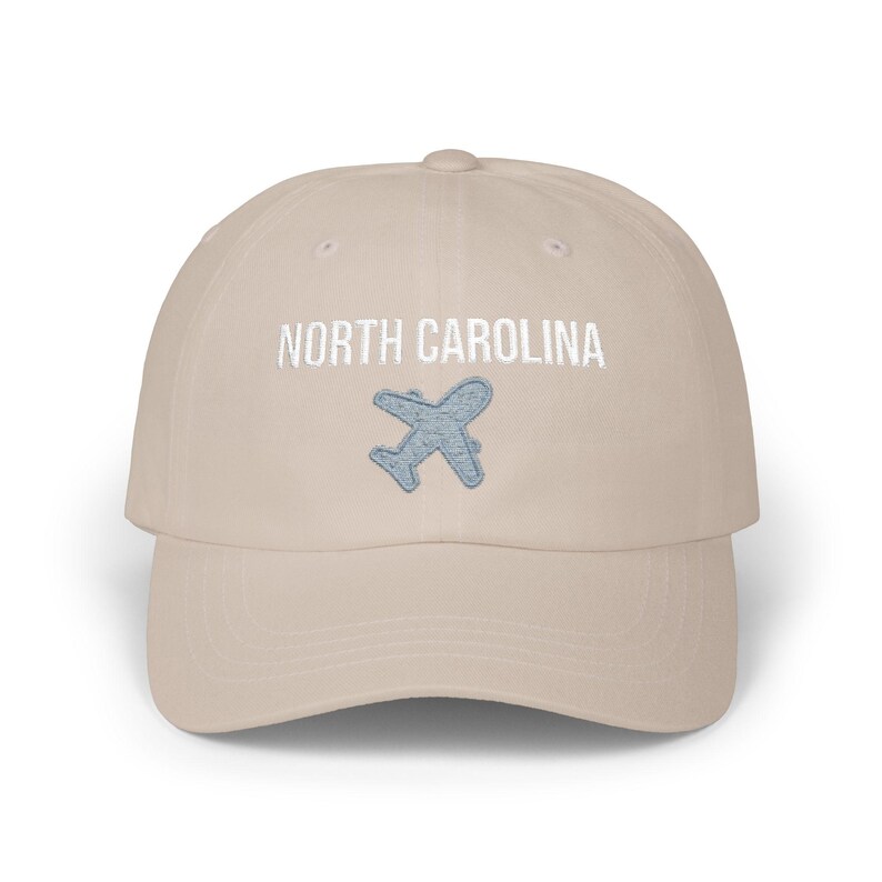 Embroidered dad cap with the words “NORTH CAROLINA” above a small light blue airplane graphic centered on the front; one-size-fits-all cotton twill hat with a curved visor and adjustable strap, available in the same color chart options as the other state caps (pink, white, black, light blue, khaki, stone, green camo, dark grey, spruce, and navy).