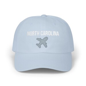 Embroidered dad cap with the words “NORTH CAROLINA” above a small light blue airplane graphic centered on the front; one-size-fits-all cotton twill hat with a curved visor and adjustable strap, available in the same color chart options as the other state caps (pink, white, black, light blue, khaki, stone, green camo, dark grey, spruce, and navy).