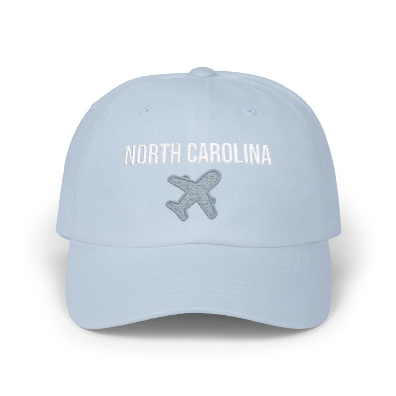 Embroidered dad cap with the words “NORTH CAROLINA” above a small light blue airplane graphic centered on the front; one-size-fits-all cotton twill hat with a curved visor and adjustable strap, available in the same color chart options as the other state caps (pink, white, black, light blue, khaki, stone, green camo, dark grey, spruce, and navy).