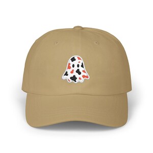 May include: A tan baseball cap featuring an embroidered ghost design. The ghost is white with black and red spots. The cap has a curved brim and a structured fit, suitable for casual wear.