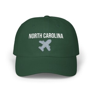 Embroidered dad cap with the words “NORTH CAROLINA” above a small light blue airplane graphic centered on the front; one-size-fits-all cotton twill hat with a curved visor and adjustable strap, available in the same color chart options as the other state caps (pink, white, black, light blue, khaki, stone, green camo, dark grey, spruce, and navy).