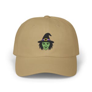 May include: A tan baseball cap featuring an embroidered witch face. The witch has green skin, black hair, and a black hat with a purple band and gold buckle. The cap is a classic six-panel design with a curved brim.