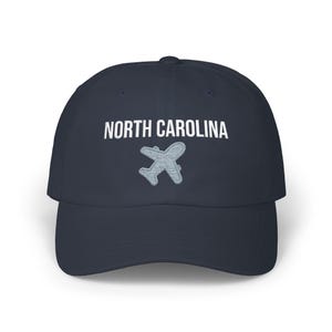 Embroidered dad cap with the words “NORTH CAROLINA” above a small light blue airplane graphic centered on the front; one-size-fits-all cotton twill hat with a curved visor and adjustable strap, available in the same color chart options as the other state caps (pink, white, black, light blue, khaki, stone, green camo, dark grey, spruce, and navy).