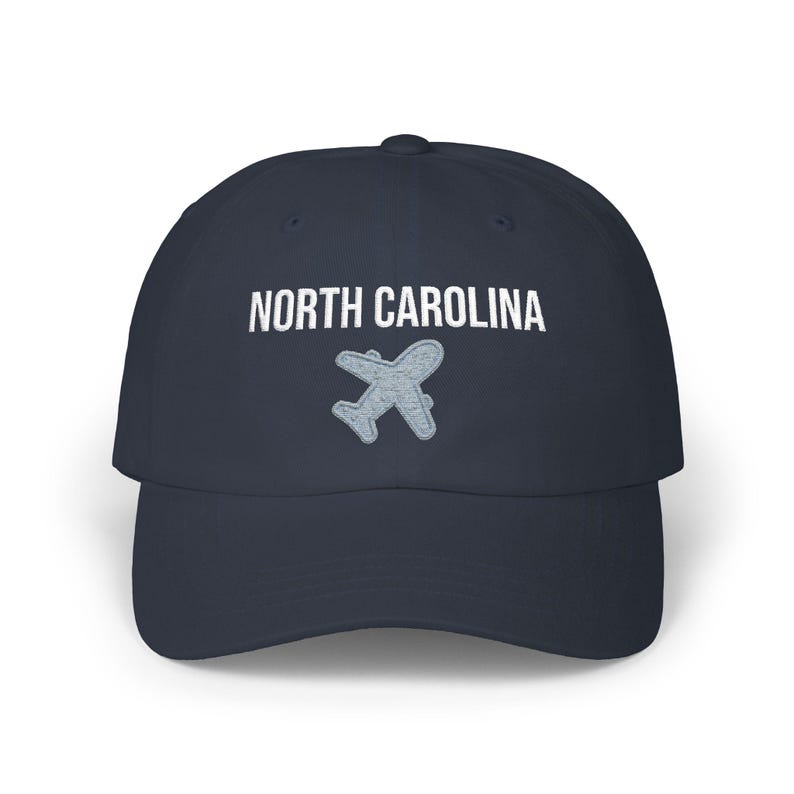 Embroidered dad cap with the words “NORTH CAROLINA” above a small light blue airplane graphic centered on the front; one-size-fits-all cotton twill hat with a curved visor and adjustable strap, available in the same color chart options as the other state caps (pink, white, black, light blue, khaki, stone, green camo, dark grey, spruce, and navy).