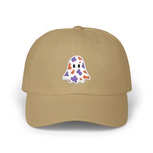 May include: A tan baseball cap featuring an embroidered ghost design. The ghost is white with purple and orange spots. The cap has a curved brim and a structured fit, suitable for casual wear.