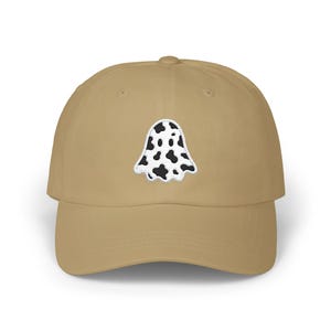 May include: A tan baseball cap with a white and black ghost-shaped patch on the front. The patch has a white border and a black and white spotted design. The cap has a curved brim and a fabric strap closure.