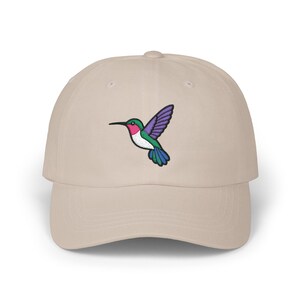 May include: A beige baseball cap featuring an embroidered hummingbird design. The hummingbird has a green head, pink throat, white chest, purple wings, and blue tail. The cap has a curved brim and a fabric strap closure.