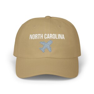 Embroidered dad cap with the words “NORTH CAROLINA” above a small light blue airplane graphic centered on the front; one-size-fits-all cotton twill hat with a curved visor and adjustable strap, available in the same color chart options as the other state caps (pink, white, black, light blue, khaki, stone, green camo, dark grey, spruce, and navy).