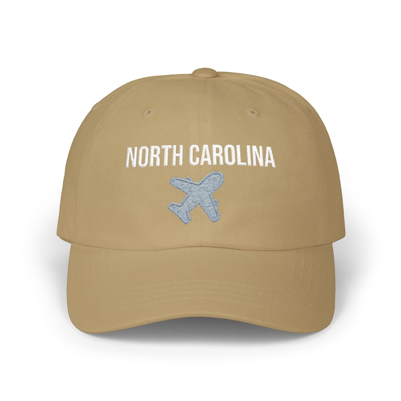 Embroidered dad cap with the words “NORTH CAROLINA” above a small light blue airplane graphic centered on the front; one-size-fits-all cotton twill hat with a curved visor and adjustable strap, available in the same color chart options as the other state caps (pink, white, black, light blue, khaki, stone, green camo, dark grey, spruce, and navy).