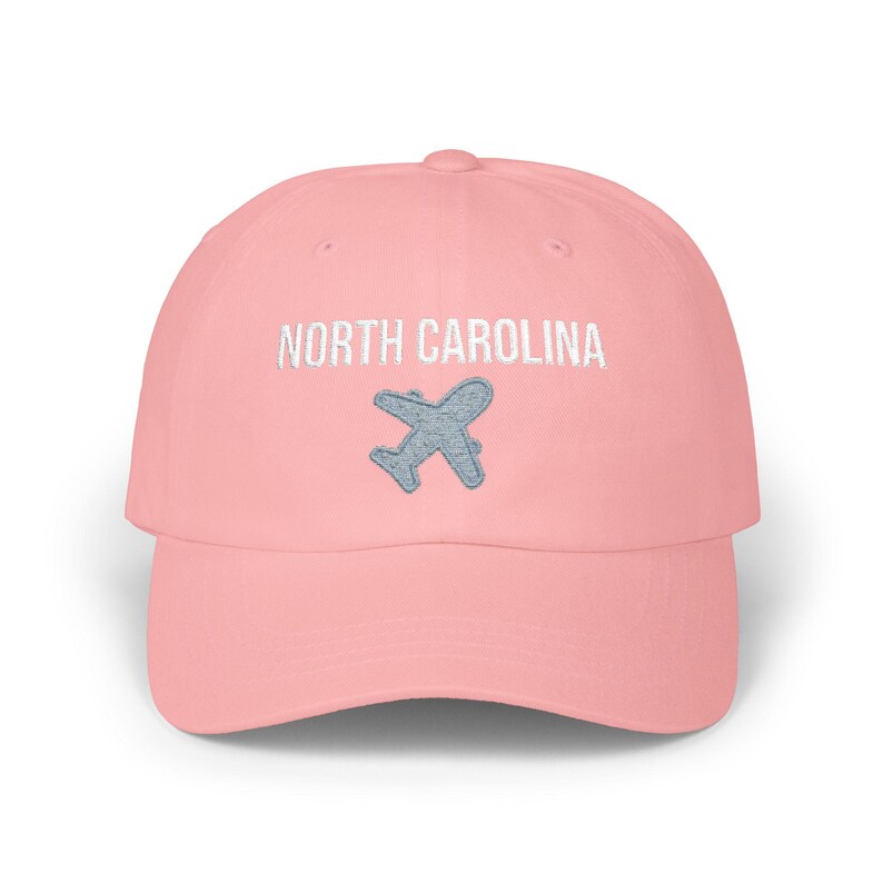 Embroidered dad cap with the words “NORTH CAROLINA” above a small light blue airplane graphic centered on the front; one-size-fits-all cotton twill hat with a curved visor and adjustable strap, available in the same color chart options as the other state caps (pink, white, black, light blue, khaki, stone, green camo, dark grey, spruce, and navy).