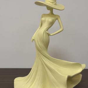 Pode incluir: Estatueta amarelo pálido de uma mulher em um vestido esvoaçante e um chapéu de abas largas. A figura tem uma mão na anca, e o vestido parece estar ao vento. O chapéu tem uma faixa decorativa ao redor da coroa. A estatueta está em uma superfície marrom escura.