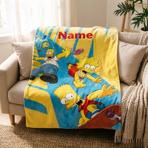 Op de afbeelding: Gele en blauwe fleece deken met stripfiguren. De deken heeft een blauwe achtergrond met gele accenten en het woord "Name" in het rood. De personages zijn onder meer Homer, Lisa en Bart Simpson.