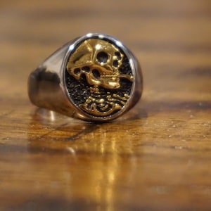 Op de afbeelding: Een zilverkleurige ring met een goudkleurig schedelontwerp. De schedel is gedetailleerd met oogkassen, een neus en tanden. De ring heeft een gepolijste afwerking en ligt op een houten oppervlak.