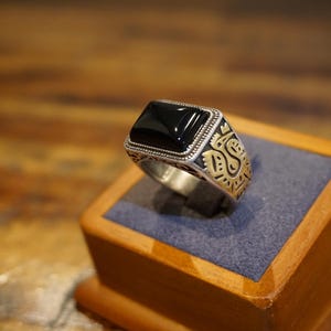 May include: A silver ring featuring a rectangular black stone set in a beaded frame. The sides of the ring band are decorated with gold-toned metal accents displaying an engraved pattern of a stylized bird or creature on both sides.