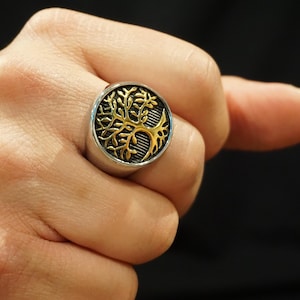 Op de afbeelding: Een zilveren ring met een cirkelvormige voorkant met een goudkleurig boomontwerp. De boom heeft ingewikkelde takken en is geplaatst tegen een zwarte achtergrond. De ring wordt aan een vinger gedragen.