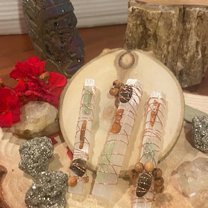 May include: Three selenite wands wrapped with copper wire and adorned with crystals and wooden beads, displayed on a wooden slice. Additional crystals, including pyrite and amethyst, are scattered around the wands. A carved decorative piece and red flowers are in the background.