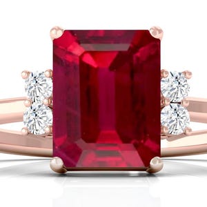 Könnte beinhalten: Roségoldener Ring mit einem großen, rechteckigen, tiefroten Rubin als Mittelstein. Zwei kleinere, runde, klare Edelsteine flankieren den Rubin auf jeder Seite. Das Band ist aus zartem Roségold.