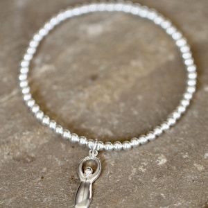 May include: A silver bracelet with a small silver charm in the shape of a woman. The bracelet is made of small silver beads.