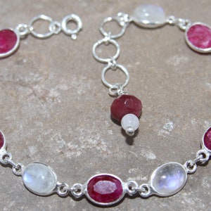 May include: A silver bracelet featuring alternating oval-shaped moonstone and ruby gemstones. The bracelet has a lobster clasp fastening and a dangling ruby charm.
