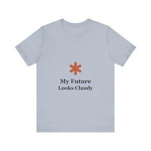 May include: A light blue t-shirt with a rust-colored asterisk graphic and the text "My Future Looks Claudy" in black. The shirt is a short-sleeved crew neck.