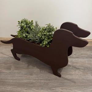 May include: A brown wooden dachshund-shaped planter filled with green leafy plants. The planter is designed with the dog's body, legs, and head, creating a unique and decorative piece for indoor or outdoor use.