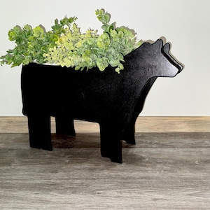 May include: A black wooden cow-shaped planter filled with green faux foliage. The planter has a cutout design and is standing on four legs. The cow planter is a decorative item for home decor.