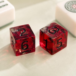 May include: Two translucent red dice with embedded gold-colored flakes and black engraved numbers. The left die shows the numbers 2, 6, and 8, while the right die displays the numbers 3, 1, and 5. The dice are square-shaped with rounded edges.