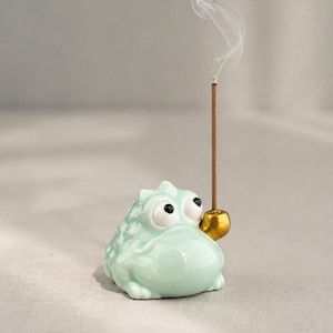 May include: A light green ceramic incense holder shaped like a cartoon frog with large eyes. The frog holds a gold-colored incense holder, and a lit incense stick emits a wisp of smoke. The holder is on a neutral background.