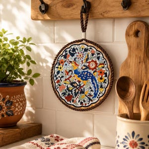 May include: A round ceramic trivet featuring a colorful hand-painted design of a bird surrounded by intricate floral patterns. The trivet is framed by a dark twisted rope border and includes a matching rope loop at the top for hanging display.