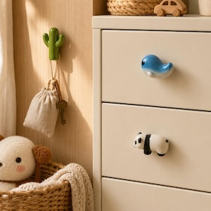 May include: A green cactus wall hook holding a beige drawstring pouch and metal keys, alongside a cream-colored dresser featuring a blue whale-shaped drawer knob and a black and white panda-shaped drawer knob on the front panels.