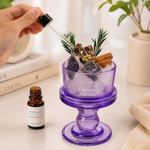 May include: A purple glass pedestal bowl filled with clear and dark purple crystals, dried eucalyptus pods, and cinnamon sticks. The bowl is labeled "Crystal Aroma" and "Oil Diffuser." A small brown bottle of Jasmine White Tea essential oil is nearby.
