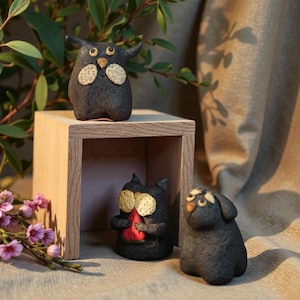 May include: Handmade ceramic figurines in a whimsical style. The set includes a dark gray owl-like creature with horns, a black cat holding a red watermelon slice, and a gray dog. The figurines have tan and beige accents.