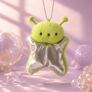 May include: A plush lime green creature keychain with a fuzzy texture, two antennae, and a small embroidered face. The body features a metallic silver star-shaped overlay attached to the front, suspended by a silver-toned metal ball chain.