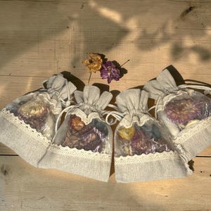 May include: Four small, gray linen drawstring bags with dried flowers visible through a clear panel. Each bag is trimmed with white lace. The bags are arranged on a wooden surface, with dried flowers in the background.