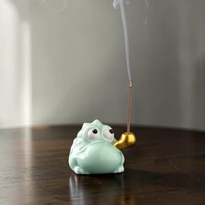 May include: A light green ceramic frog incense holder with a gold-colored pipe. The frog has large, expressive eyes and is holding an incense stick that is emitting smoke. The holder is on a dark brown surface.