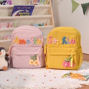 May include: A light pink corduroy backpack with the name Amelia in colorful letters and a yellow flower charm, and a yellow corduroy backpack with the name Sophia in colorful letters and a pink flower charm, both featuring front pockets.