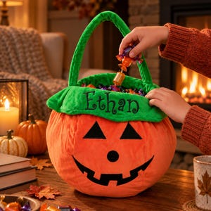 May include: A plush orange pumpkin-shaped Halloween candy bucket with a green fabric handle and rim. The front features a black felt jack-o'-lantern face and the name Ethan embroidered in black thread on the green rim, filled with assorted candies.