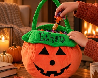 May include: A plush orange pumpkin-shaped treat bucket with a green fabric rim embroidered with the name Ethan in black thread. The bucket features a black felt jack-o'-lantern face with triangular eyes, a round nose, and a wide, jagged grin.