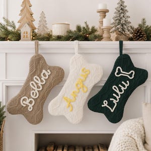 May include: Three bone-shaped Christmas stockings made of cable-knit fabric. The left stocking is tan with the white name Bella, the center is white with the yellow name Angus, and the right is dark green with the white name Lulu and a bone icon.