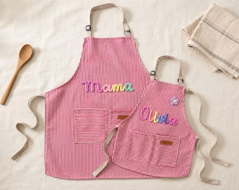May include: Two red and white pinstriped aprons with adjustable beige neck straps and front pockets. The larger apron features the word Mama in colourful puffy letters, while the smaller apron displays the name Olivia and a small purple flower.