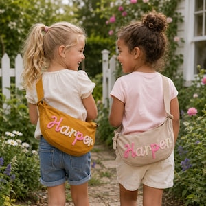 May include: Two crescent-shaped crossbody bags with adjustable straps. One bag is mustard yellow with the name Harper embroidered in pink and white. The other bag is beige with the name Harper embroidered in pink and beige. Both bags feature a zipper.